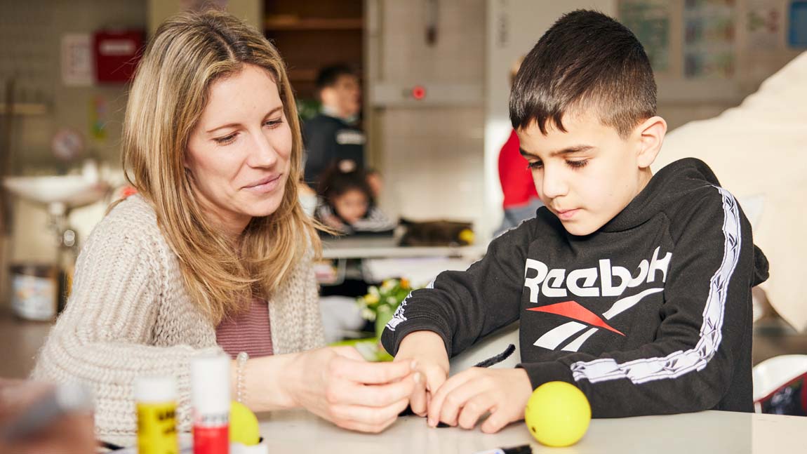 Eine Grundschullehrerin ist neben ihrem Schüler an seinem Tisch in der Hocke und hilft beim Basteln