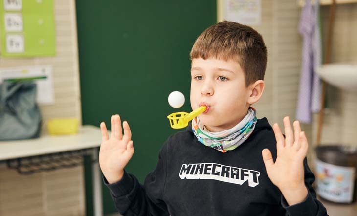 Grundschulkind sitzt im Klassenzimmer und pustet mit Atemtrainer Ball in die Luft