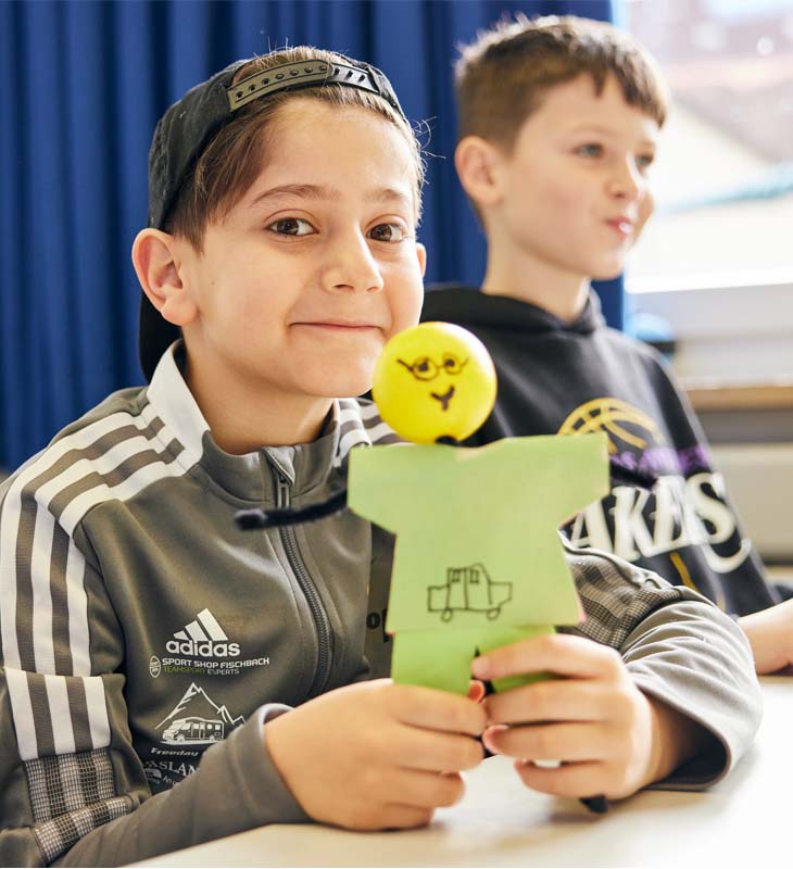 lächelnder Junge sitzt am Tisch im Klassenzimmer und hält KLARO in der Hand