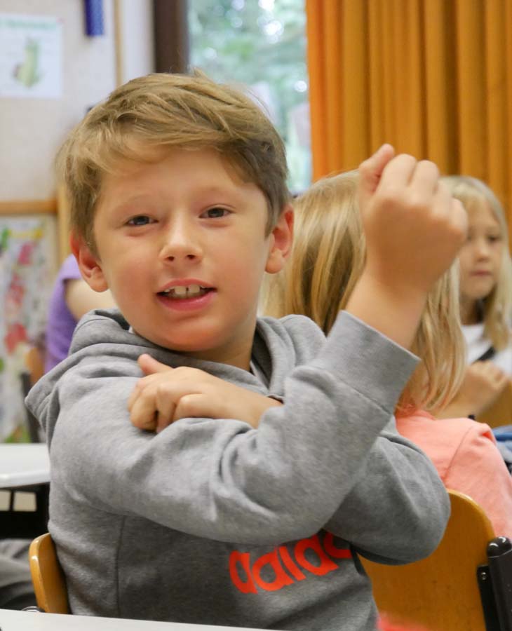 Ein blonder Junge im Grundschulalter sitzt im Klassenzimmer, blickt lächelnd in die Kamera und fühlt seinen angespannten Bizeps. 