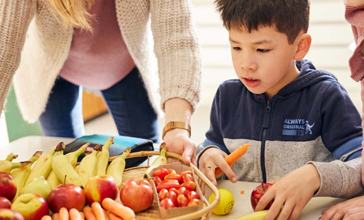 Eine Frau reicht einem dunkelhaarigen Jungen ein Tablett mit Bananen, Tomaten, Karotten und Äpfeln.