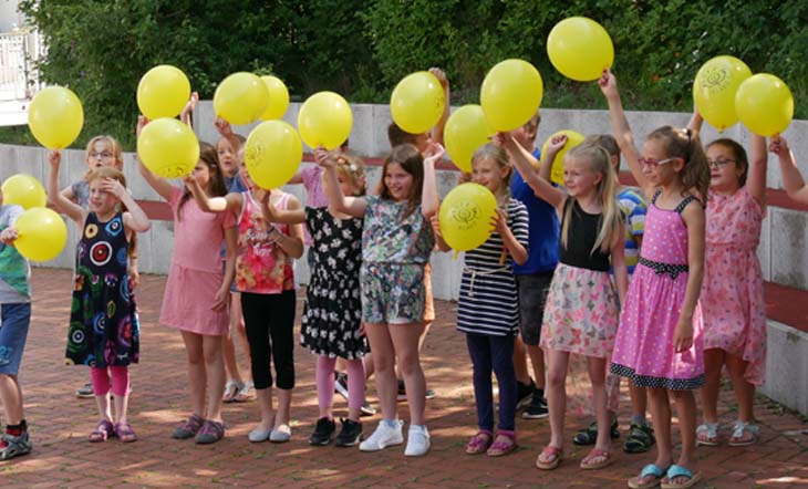 Eine Gruppe von Grundschulkindern steht fröhlich in einem Pausenhof und winkt ausgelassen mit gelben Luftballons. 