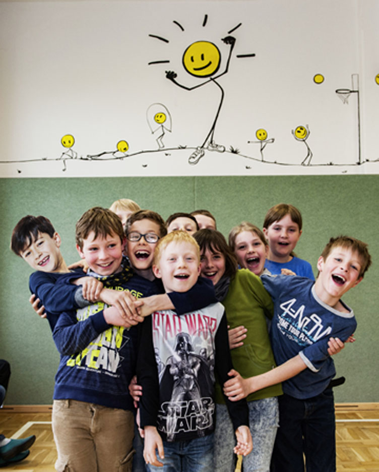 Eine Gruppe von elf lachenden Kindern im Grundschulalter steht in einer Turnhalle sehr eng zusammen. Die Kinder halten sich gegenseitig fest. Über ihnen sind sportliche KLAROs an die Wand gemalt.