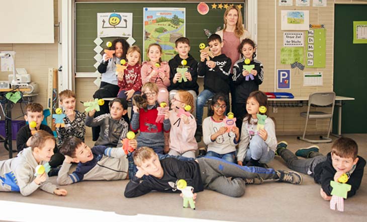 Eine Gruppe von fröhlichen 2. Klässler:innen steht gemeinsam mit der Klassenlehrerin in ihrem Klassenraum vor der Tafel und hält gebastelte KLAROs in die Kamera. Im Hintergrund sind Klasse2000 Plakate erkennbar.