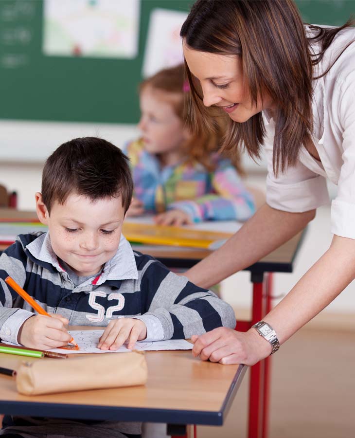 Eine junge brünette Lehrerin beugt sich zu einem sitzenden, jungen Schüler. Der Junge grinst verschmitzt und schreibt in sein vor sich liegendes Heft. 