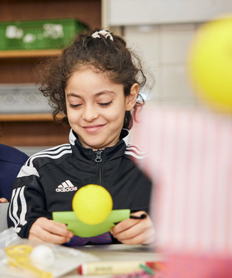 Ein Mädchen mit dunklen, lockigen Haaren sitzt im Klassenzimmer an ihrem Tisch und blickt verträumt auf ihren gebastelten KLARO.