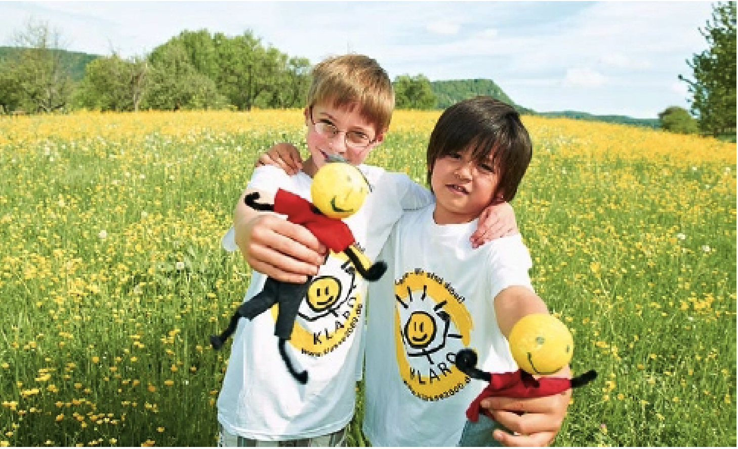 Zwei neunjährige, frech grinsende Freunde mit weißen T-Shirts umarmen sich auf einer sommerlichen Blumenwiese. Die beiden Jungen halten ihrer gebastelten KLAROs in die Kamera.