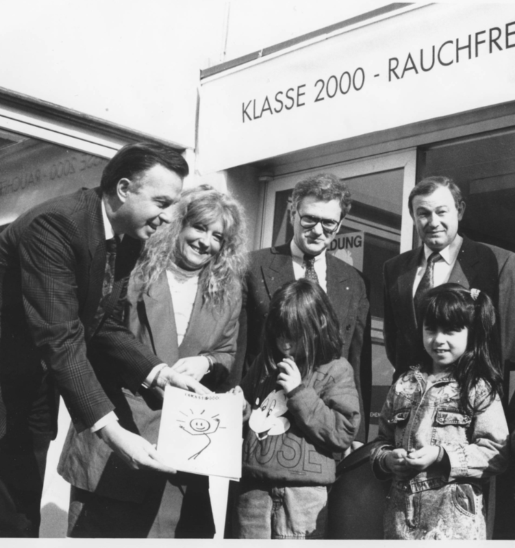 Auf einem schwarz-weiß Bild von 1991 zeigt eine Gruppe von vier Erwachsenen gemeinsam mit 2 Mädchen ein Heft "Klasse2000 - Rauchfrei" in die Kamera