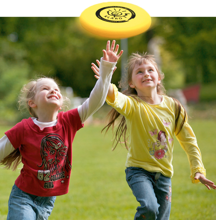 zwei zehn Jährige Mädchen laufen lachend auf einer grünen Wiese einer gelben Frisbeescheibe hinterher.