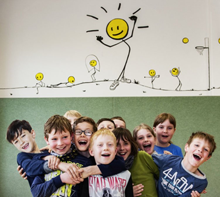 Eine Gruppe von elf lachenden Kindern im Grundschulalter steht in einer Turnhalle sehr eng zusammen. Die Kinder halten sich gegenseitig fest. Über ihnen sind sportliche KLAROs an die Wand gemalt.