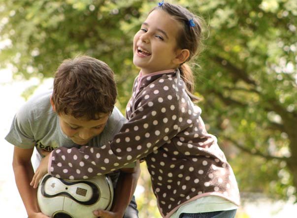 In einem Garten versucht ein lachendes Mädchen ihrem lachenden Freund einen Fußball aus den Händen zu nehmen. 