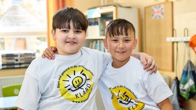 Zwei fröhliche Jungs tragen KLARO T-Shirts. Sie umarmen sich im Klassenzimmer.