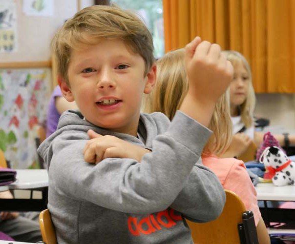 Ein blonder Junge im Grundschulalter sitzt im Klassenzimmer, lächelt in die kamera und fühlt seinen angespannten Bizeps. 